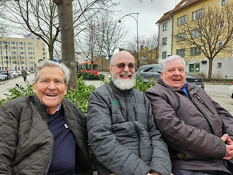 Tre glada äldre herrar sitter på en parkbänk.