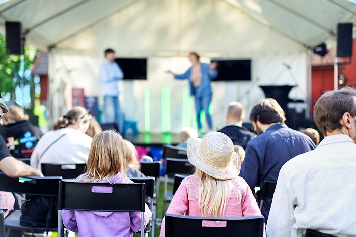 En publik med både barn och vuxna sitter på stolar framför en utomhusscen under ett tält. På scenen står två personer och pratar. I förgrunden syns ett barn med hatt som tittar mot scenen. Solen lyser och stämningen är avslappnad.
