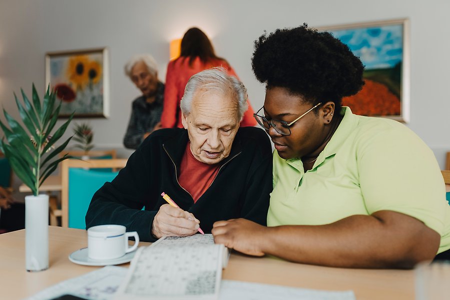 Miljö från ett äldreboende. I förgrunden en anställd sittandes med en boende vid ett bord.